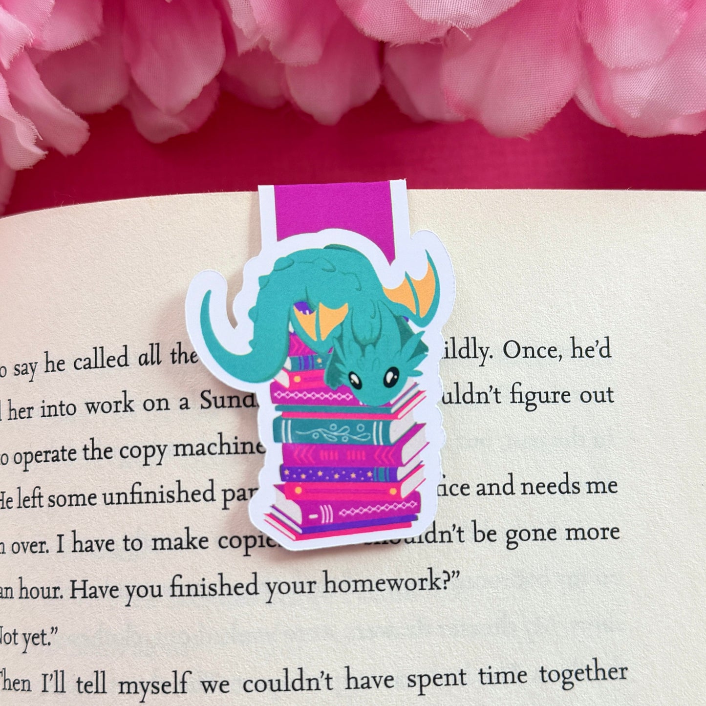 Magnetic bookmark with a teal and yellow dragon design on an open book surrounded by pink flowers.