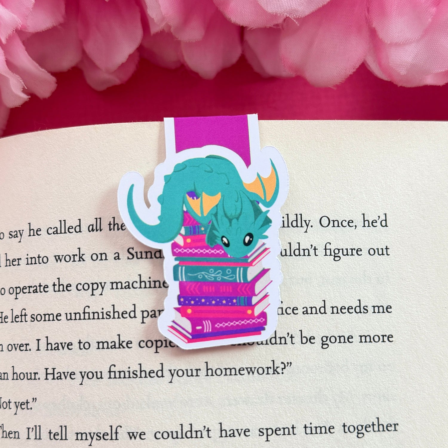 Magnetic bookmark with a teal and yellow dragon design on an open book surrounded by pink flowers.