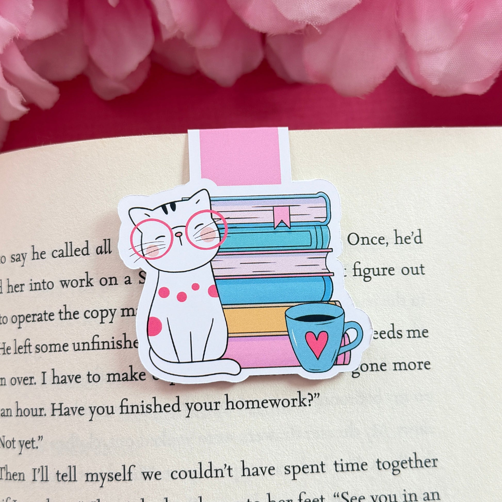 Magnetic bookmark that has a design of a cat and coffee mug sitting in front of a bookstack. The bookmark is on an open book surrounded by pink flowers.