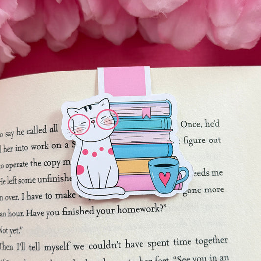 Magnetic bookmark that has a design of a cat and coffee mug sitting in front of a bookstack. The bookmark is on an open book surrounded by pink flowers.