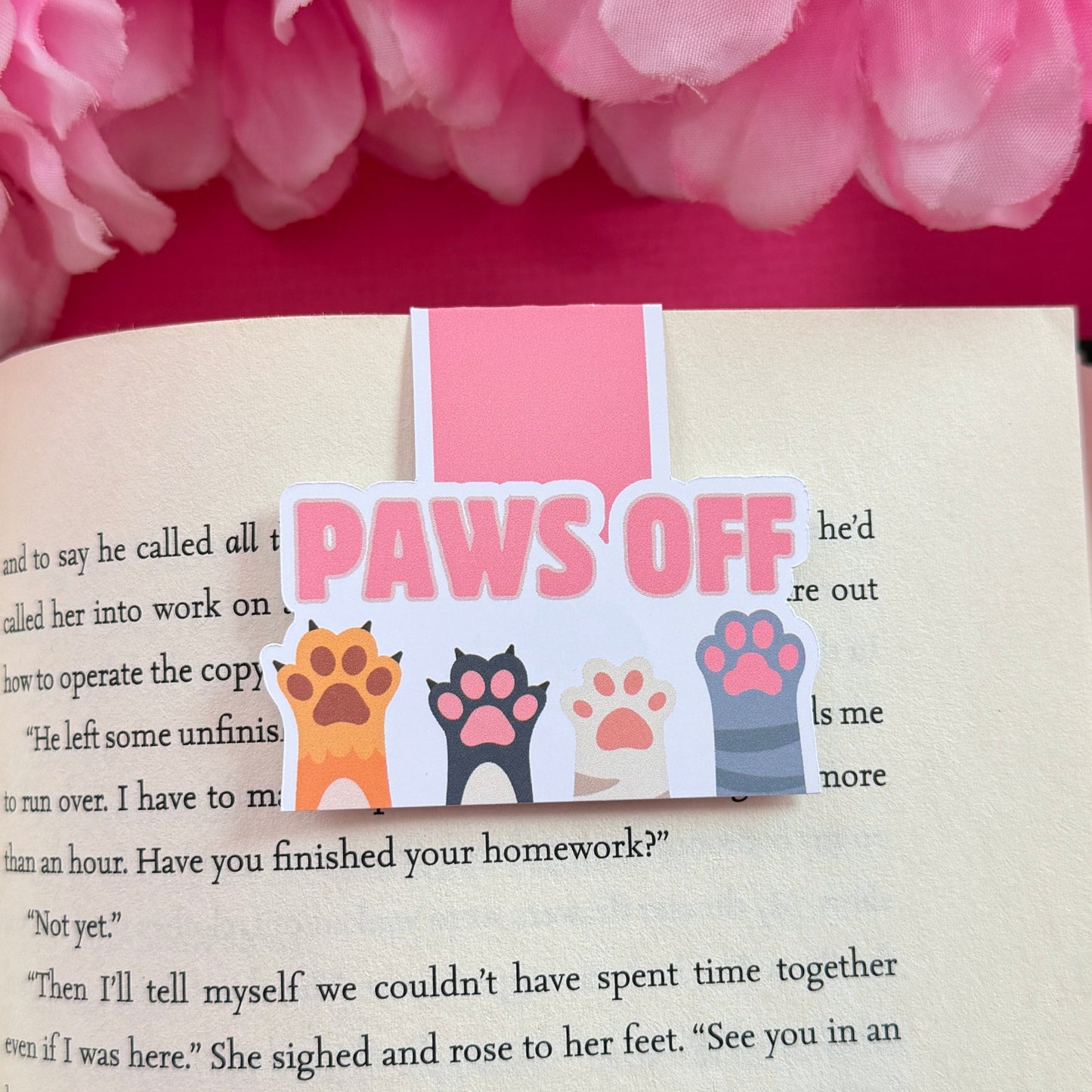 Magnetic bookmark that has a design with four cat/dog paws and the words "Paws Off". The bookmark is on an open book surrounded by pink flowers.