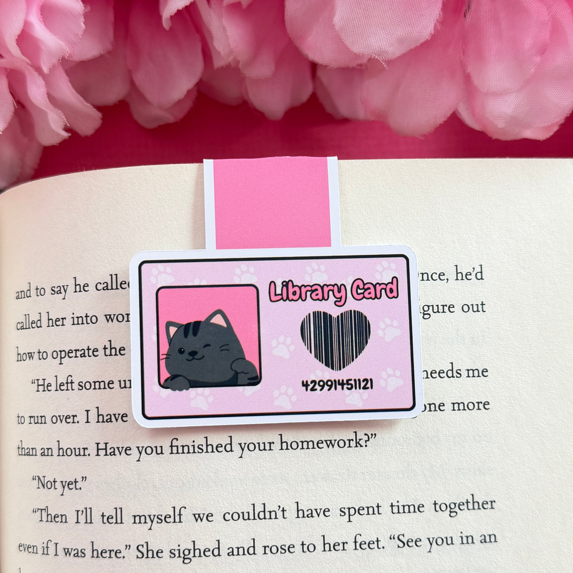 Magnetic bookmark that has a design of a pink library card with a black cat. The bookmark is on an open book surrounded by pink flowers.