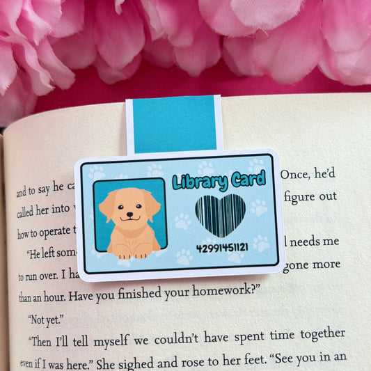 Magnetic bookmark that has a design of a blue library card with a dog. The bookmark is on an open book surrounded by pink flowers.