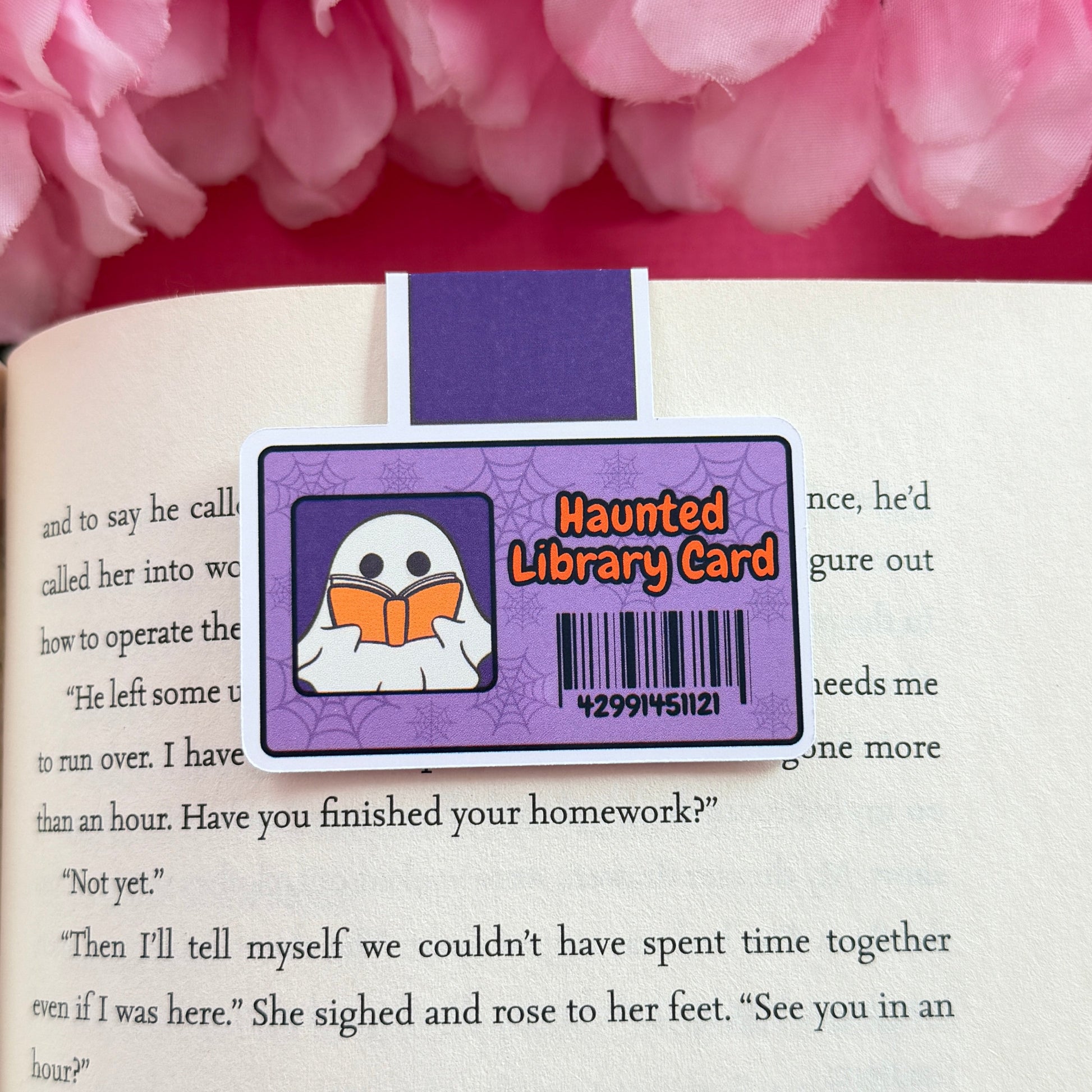 Magnetic bookmark that has a design of a haunted library card including a ghost. The bookmark is on an open book surrounded by pink flowers.