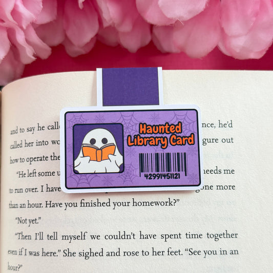 Magnetic bookmark that has a design of a haunted library card including a ghost. The bookmark is on an open book surrounded by pink flowers.