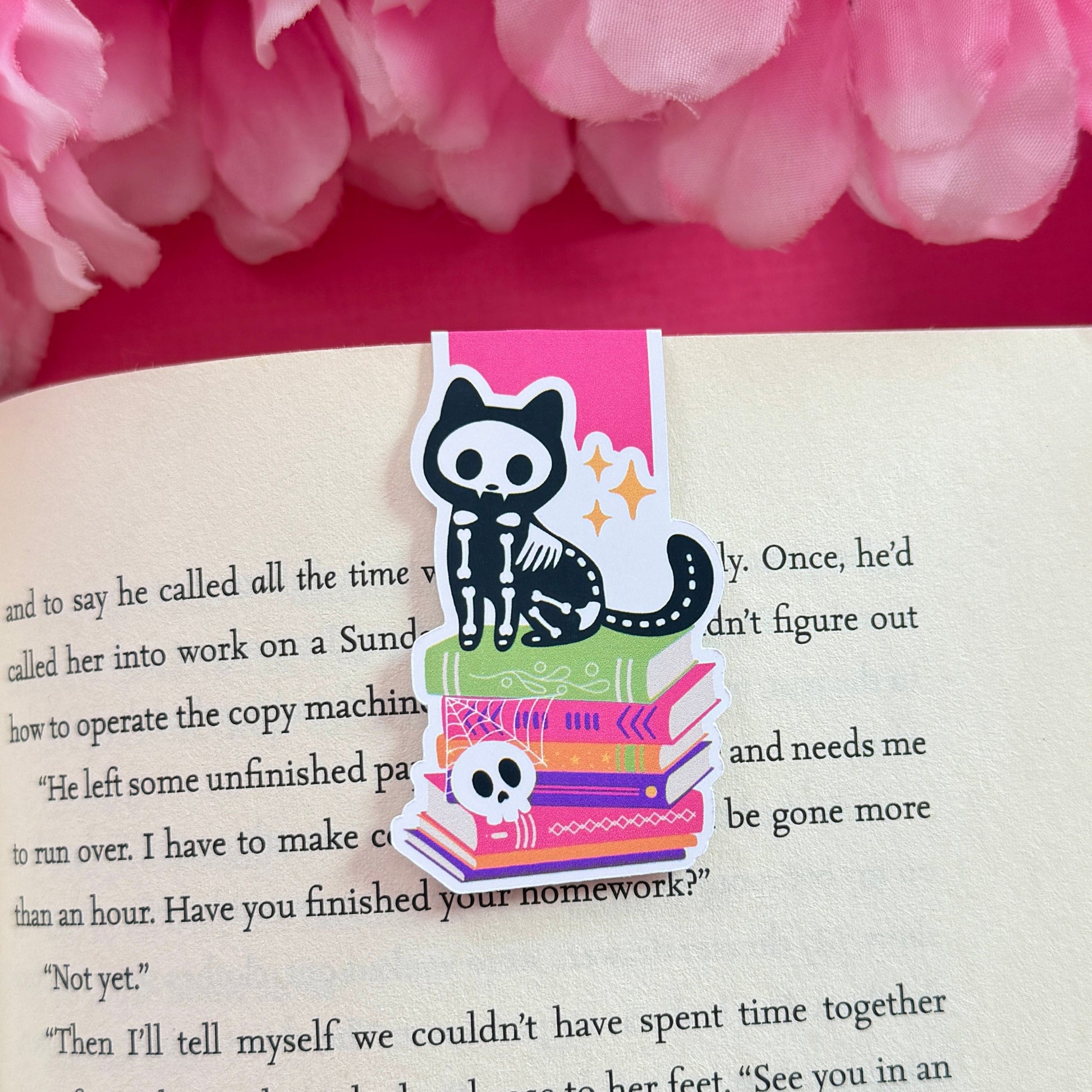 Magnetic bookmark that has a design of cute skeleton cat sitting on top of a bookstack. The bookmark is on an open book surrounded by pink flowers.