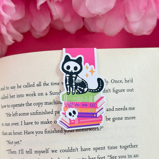 Magnetic bookmark that has a design of cute skeleton cat sitting on top of a bookstack. The bookmark is on an open book surrounded by pink flowers.