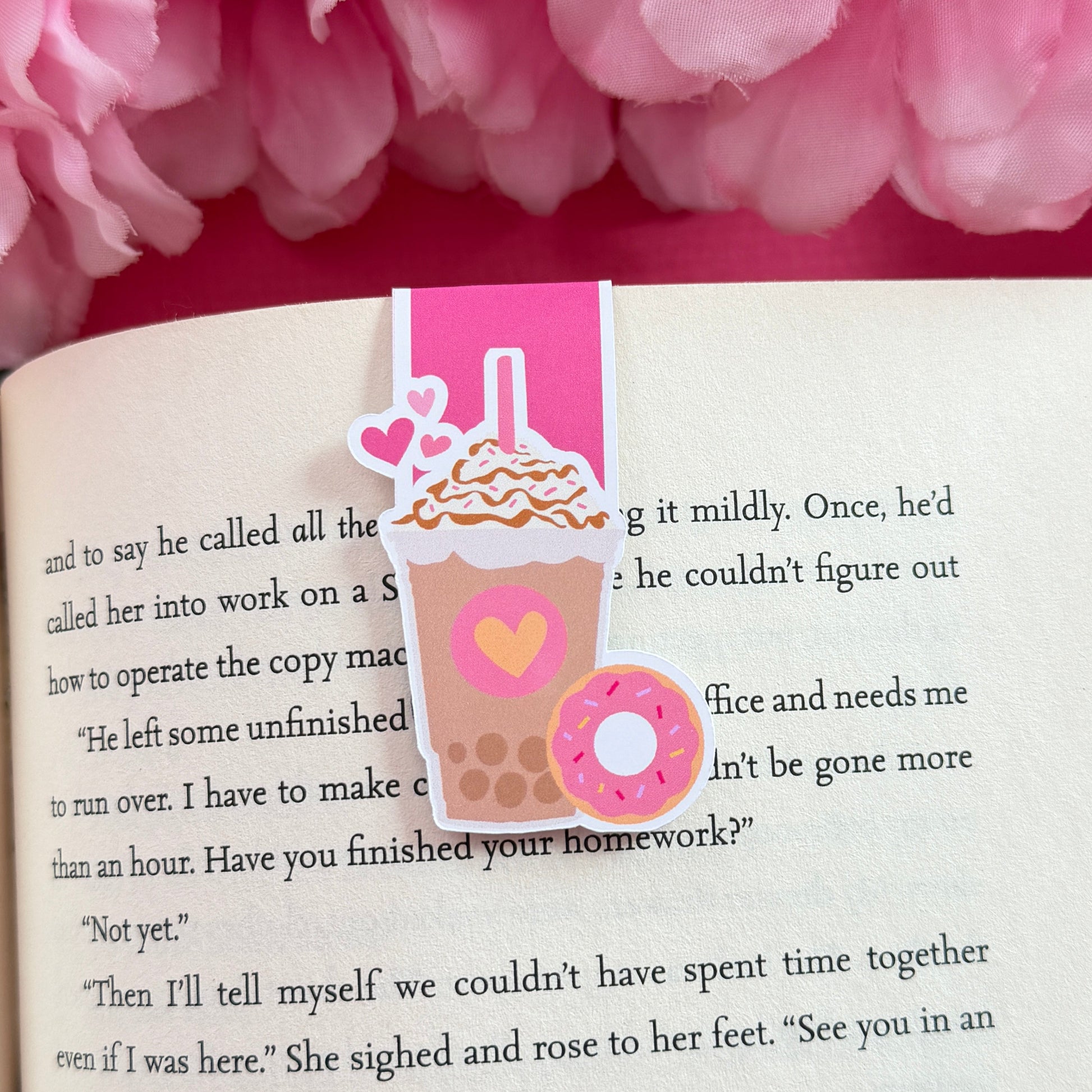 Magnetic bookmark that has a design of an iced coffee with a pink donut. The bookmark is on an open book surrounded by pink flowers.