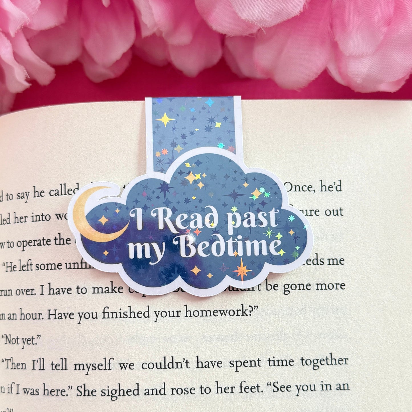 Magnetic bookmark that has a later night read design. The bookmark is on an open book surrounded by pink flowers.