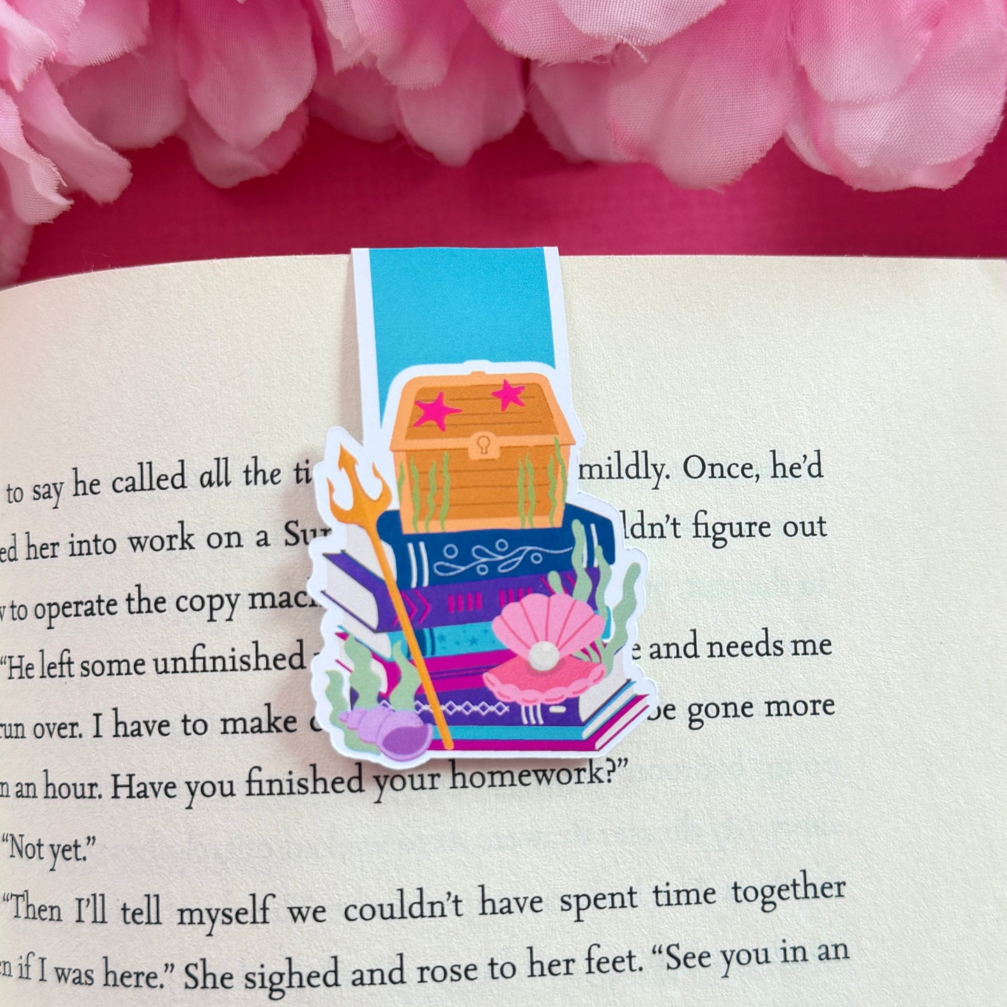 Magnetic bookmark featuring an ocean theme including a treasure chest sitting on a bookstack. The bookmark is on an open book surrounded by pink flowers.