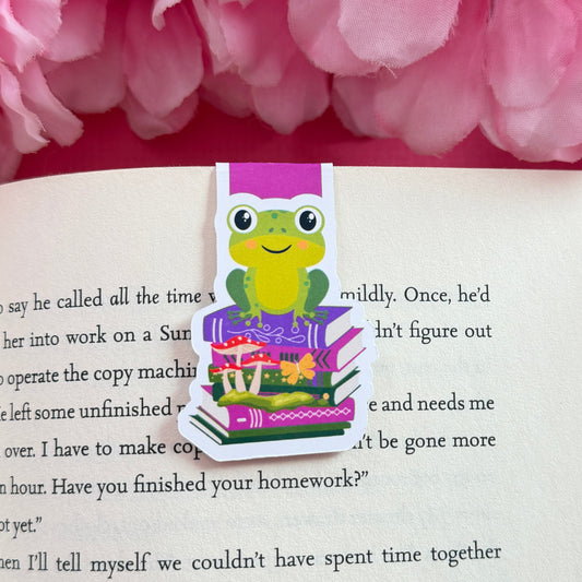 Magnetic bookmark that has a frog sitting on top of a book stack. The bookmark is on an open book surrounded by pink flowers.