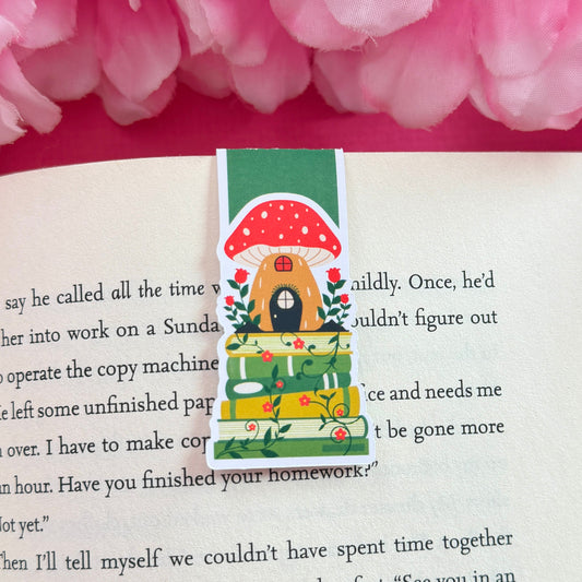 Magnetic bookmark that features a mushroom house sitting on a cottagecore-themed bookstack. The bookmark is on an open book surrounded by pink flowers.