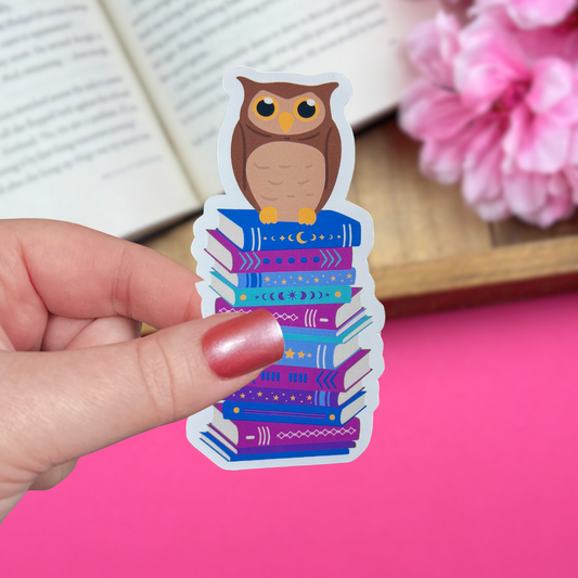 Hand holding a vinyl sticker with an owl sitting atop a late night reader themed bookstack, with an open book and pink flowers in the background.