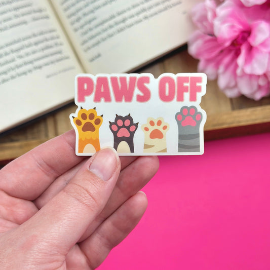 Hand holding a sticker with paw prints and 'PAWS OFF' text, with a book and flowers in the background.