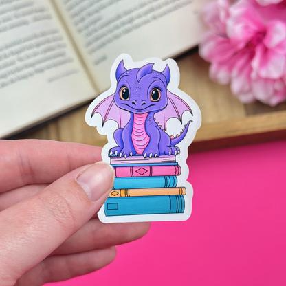 Hand holding a vinyl sticker with a purple and pink dragon sitting atop a bookstack, with an open book and pink flowers in the background.