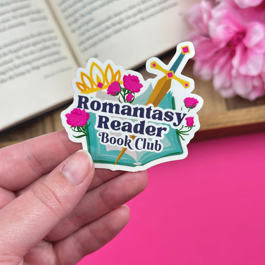Hand holding a vinyl sticker that has a fantasy design that says "romantasy reader book club", with an open book and flowers in the background.