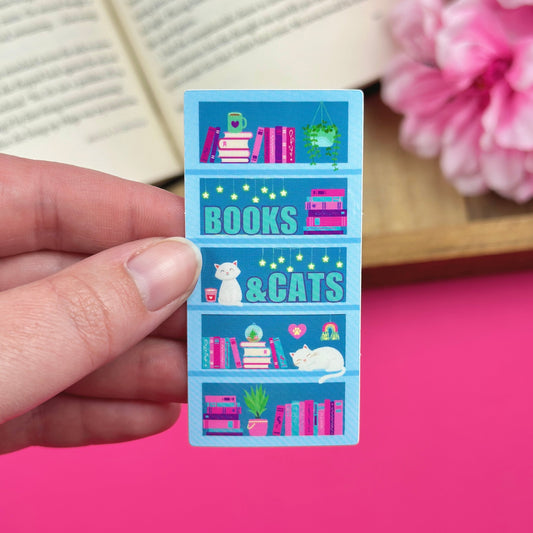 Sticker with bookshelf design featuring books and cats held by a hand in front of a pink background with an open book and flowers.