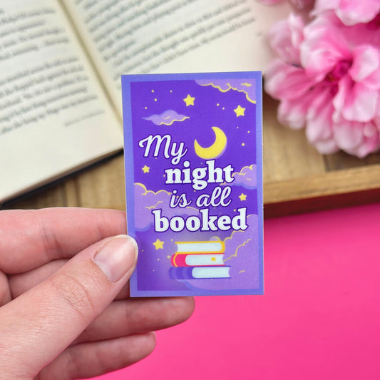 Hand holding a purple vinyl sticker that says 'My night is all booked' with a book and flowers in the background.