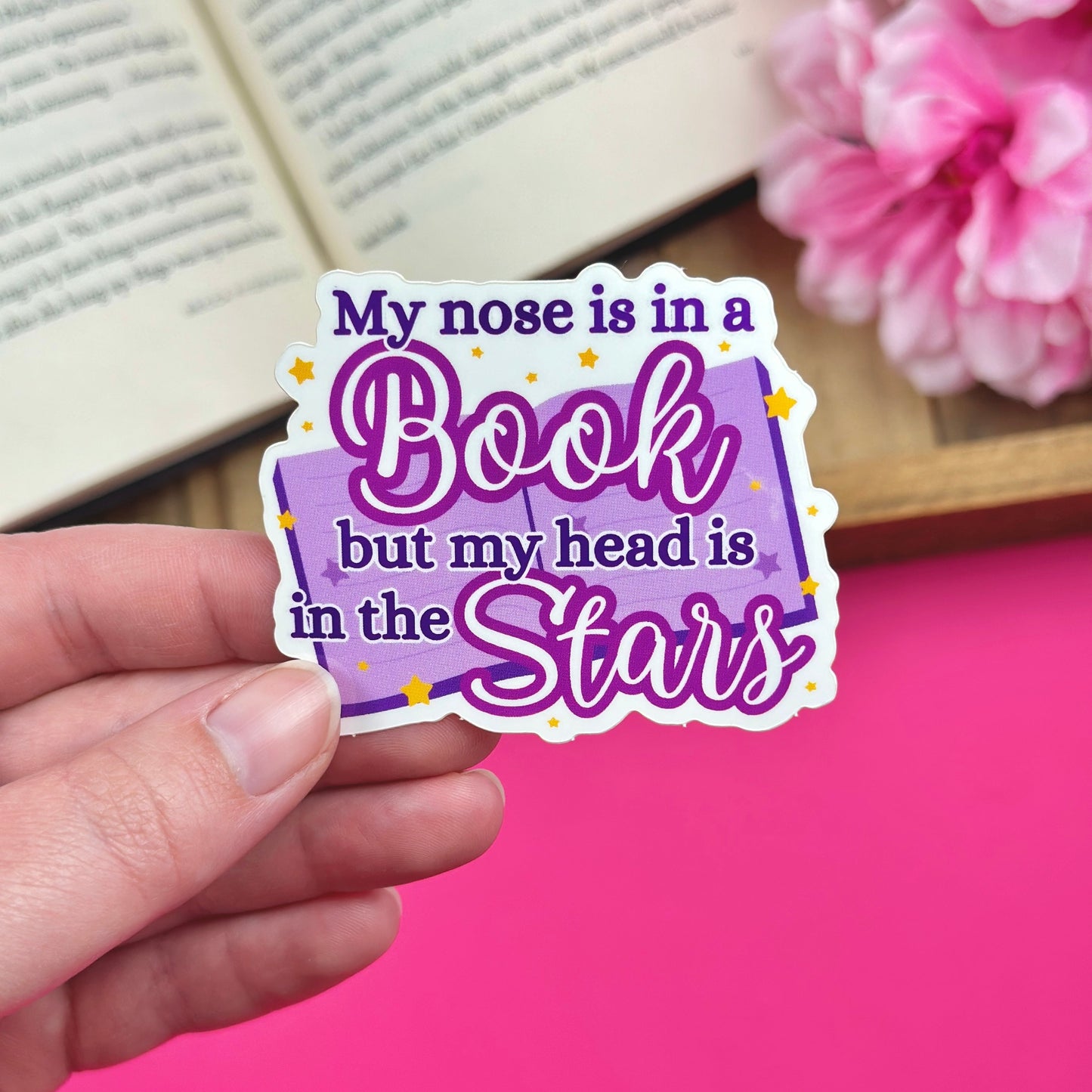 Sticker with 'My nose is in a book but my head is in the stars' text held by a hand with a pink background and open book.