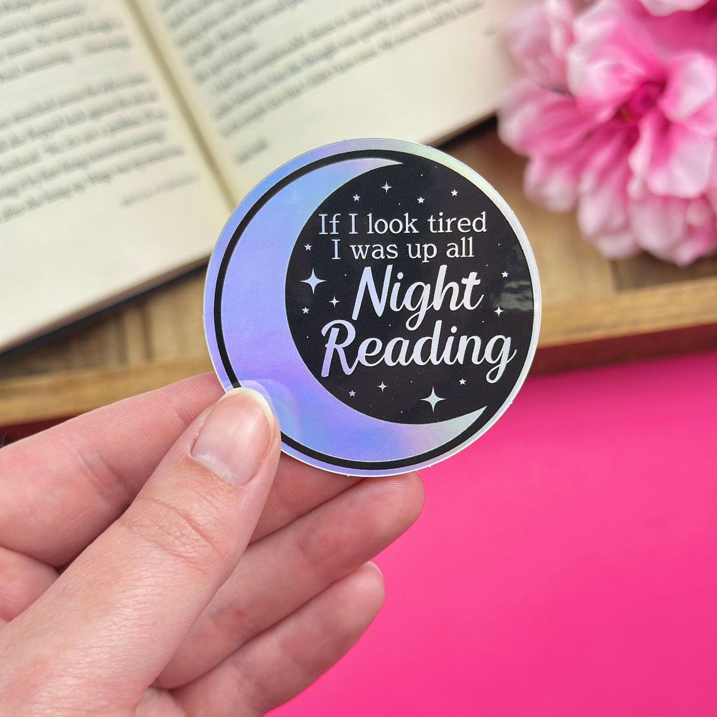 Hand holding a round sticker with text 'If I look tired, I was up all night reading' against a pink background.
