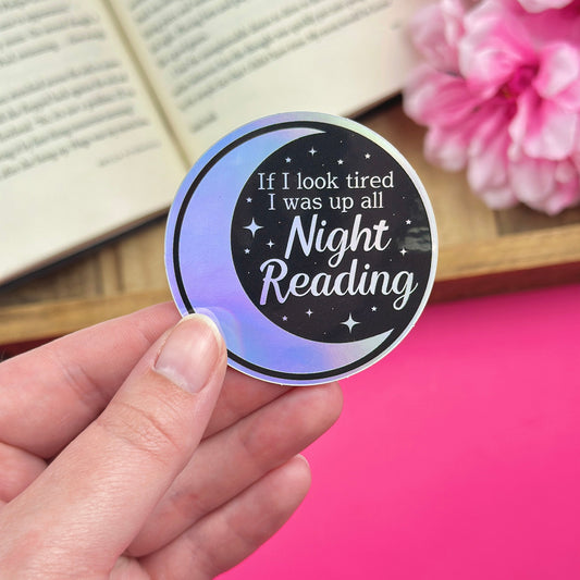 Hand holding a round sticker with text 'If I look tired, I was up all night reading' against a pink background.