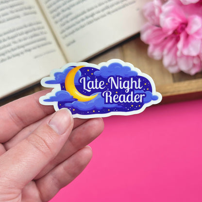 Sticker with 'Late Night Reader' design held by a hand, with flowers and an open book in the background.