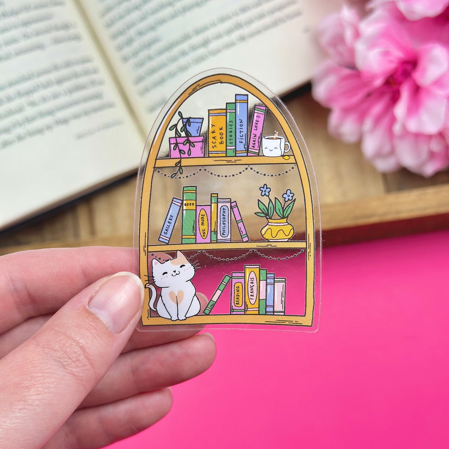 Hand holding a clear vinyl sticker with a bookshelf design, with an open book and flowers in the background.