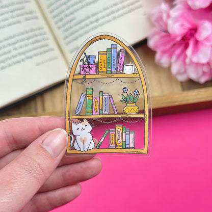Hand holding a clear vinyl sticker with a bookshelf design, with an open book and flowers in the background.