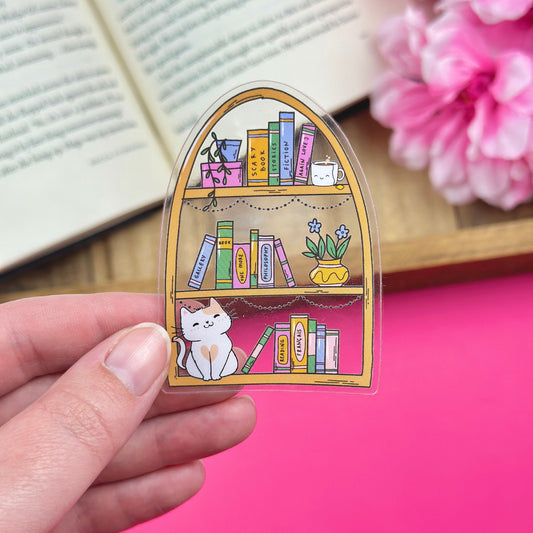 Hand holding a clear vinyl sticker with a bookshelf design, with an open book and flowers in the background.