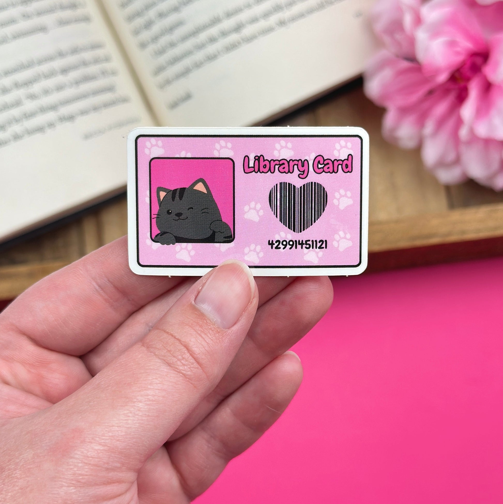 Hand holding a pink library card with a cat design with an open book and flowers in the background.
