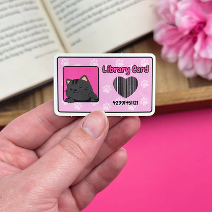 Hand holding a pink library card with a cat design with an open book and flowers in the background.