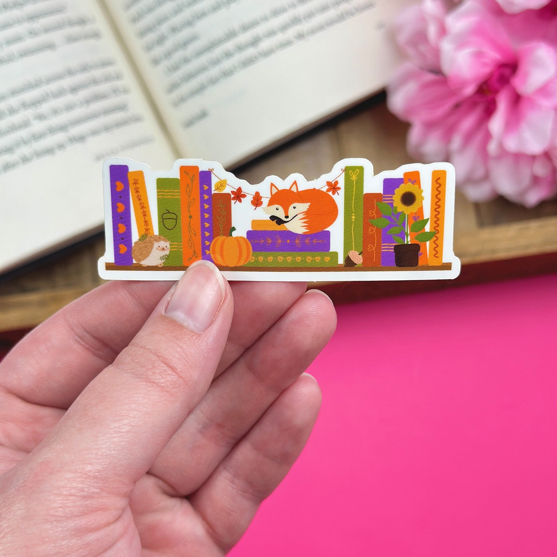 Hand holding a colorful sticker with an autumn bookshelf design that includes a fox, hedgehog, sunflower and pumpkin against a pink background.