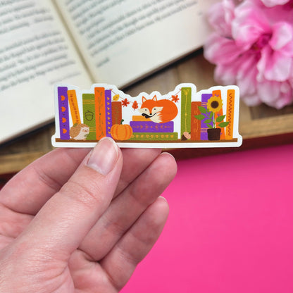 Hand holding a colorful sticker with an autumn bookshelf design that includes a fox, hedgehog, sunflower and pumpkin against a pink background.
