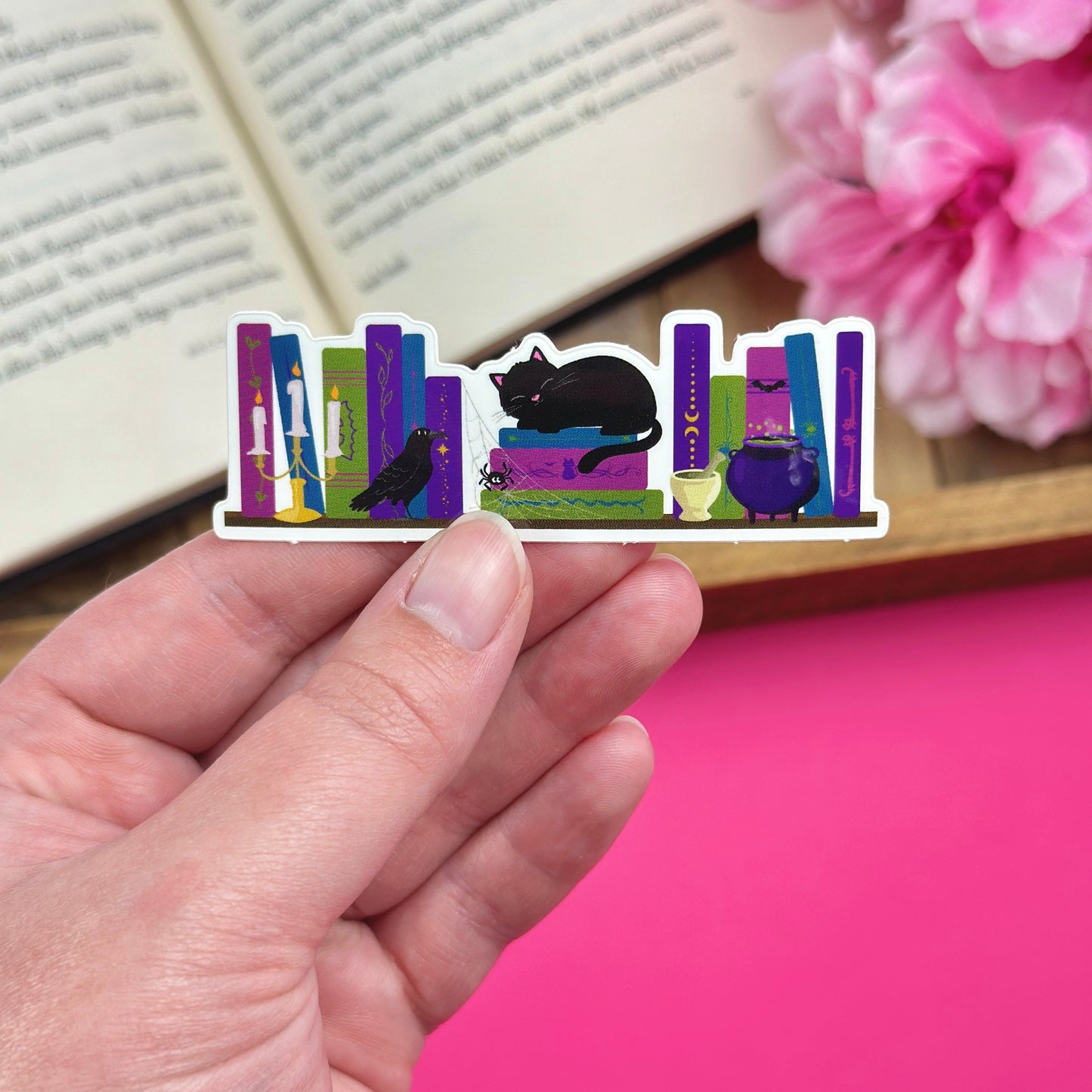 Hand holding a sticker with a dark academia bookshelf design that includes a black cat, cauldron, raven and candelabra. The sticker is being held over a pink background with an open book and pink flowers.