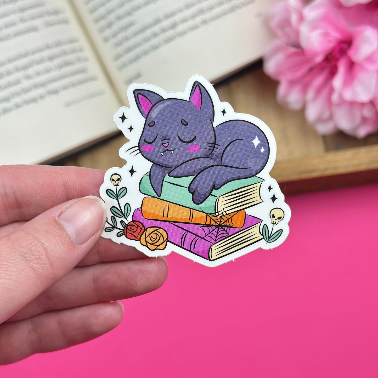 Hand holding a sticker of a black cat that is laying on a stack of books with a dark academia theme.  The sticker is being held in front of a pink background that has pink flowers and an open book laying on it.