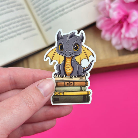 Hand holding sticker with a black and gold dragon sitting on top of bookstack, with an open book and pink flowers in the background.
