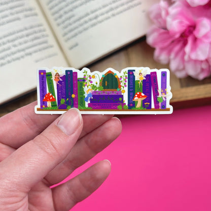 Hand holding a colorful sticker with an enchanted garden bookshelf design that includes four fairies, two mushrooms, and a fairy door against a pink background with an open book.
