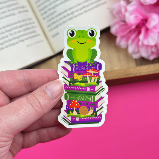 A whimsical sticker featuring a green frog perched atop a stack of purple and green books. The books are adorned with illustrations of mushrooms, a snail, and a butterfly. The frog has large, friendly eyes and a smiling expression.