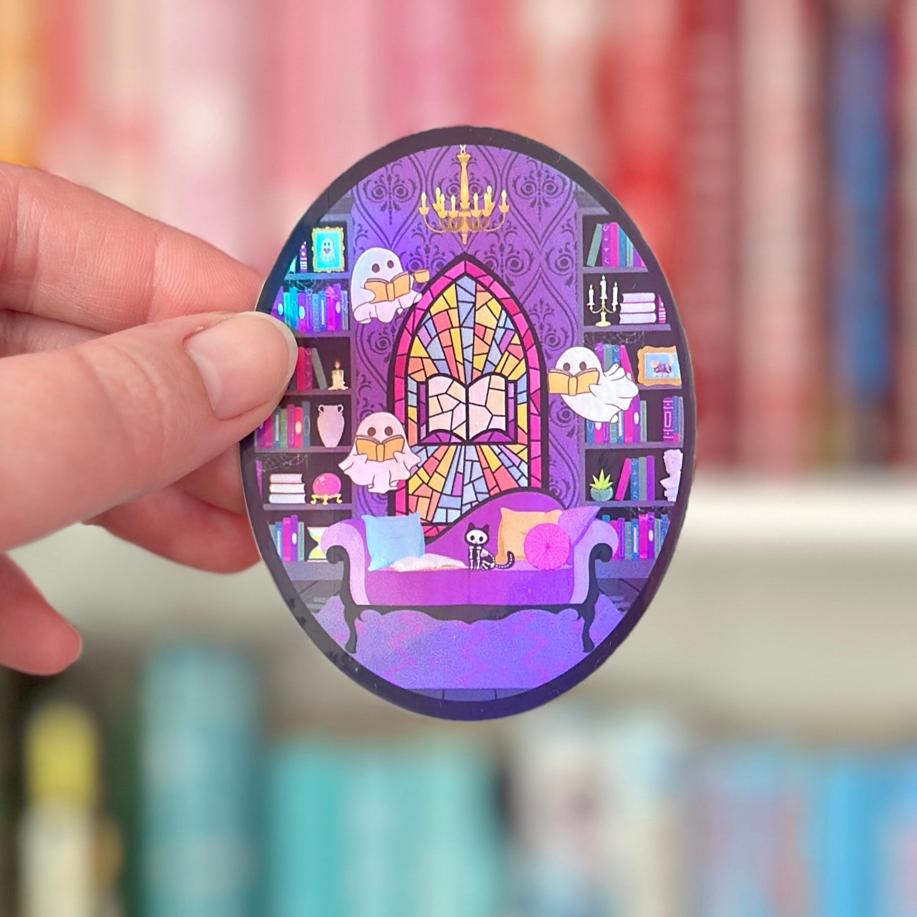 Oval-shaped sticker with a haunted library-themed design held by a hand against a blurred bookshelf background.