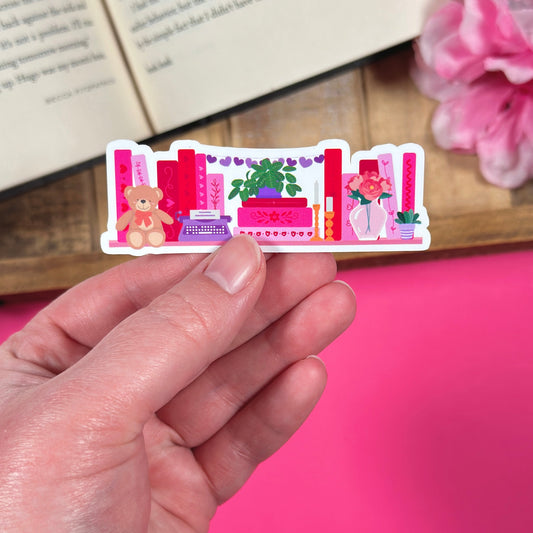 Sticker with 'romance reader bookshelf' design held by a hand, with flowers and an open book in the background.