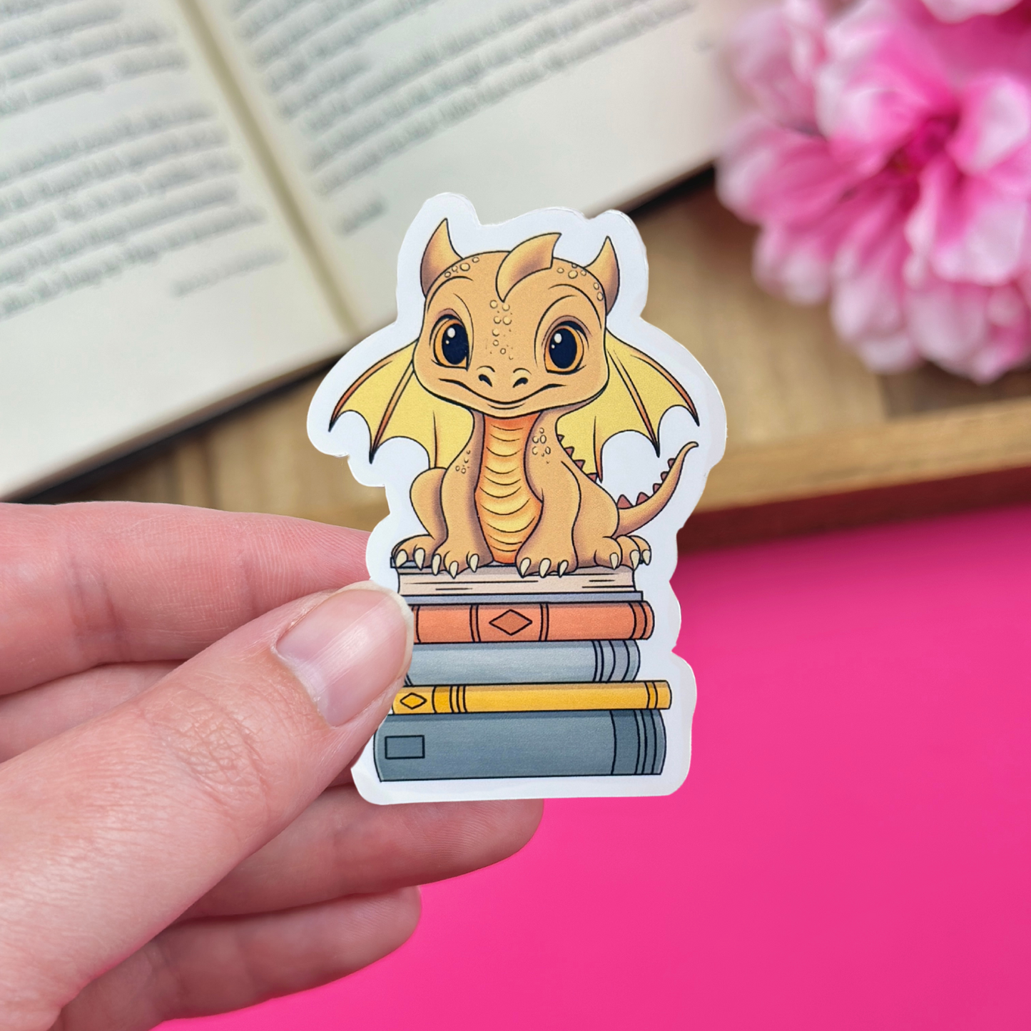 Hand holding a vinyl sticker with a golden yellow dragon sitting atop a bookstack, with an open book and pink flowers in the background.