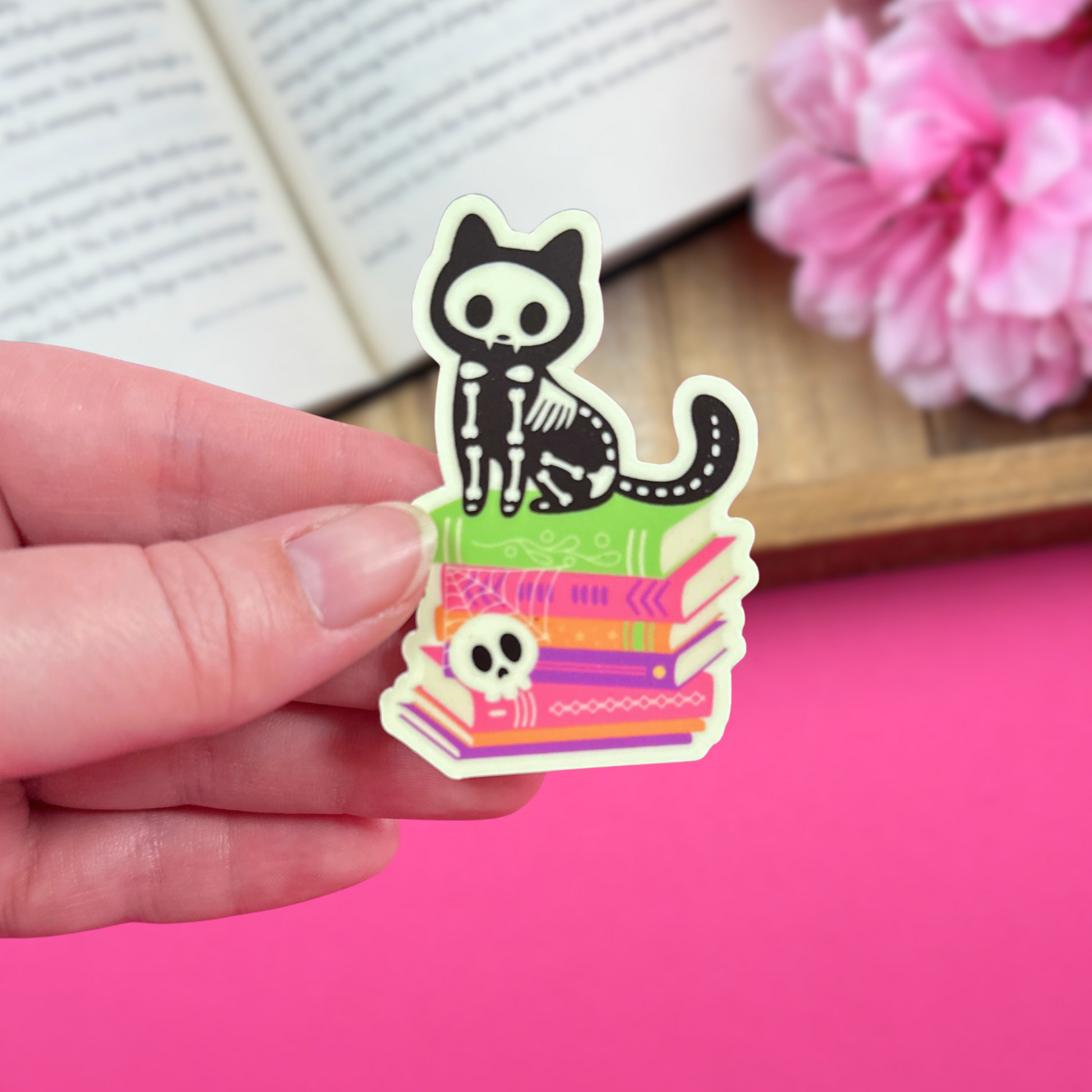 Hand holding a vinyl sticker with a skeleton cat sitting on a Halloween-themed bookstack, with an open book and pink flowers in the background. The sticker is made of matte vinyl which glows in the dark.