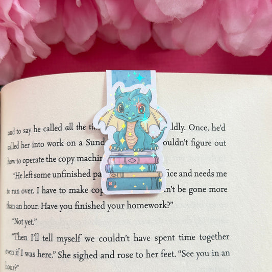 Magnetic bookmark with a teal and yellow dragon design on an open book surrounded by pink flowers. The bookmark has a holographic star finish.