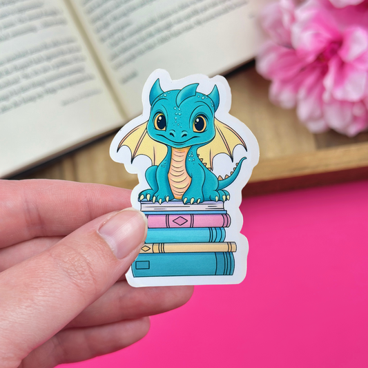 Hand holding a vinyl sticker with a teal and yellow dragon sitting atop a bookstack, with an open book and pink flowers in the background.