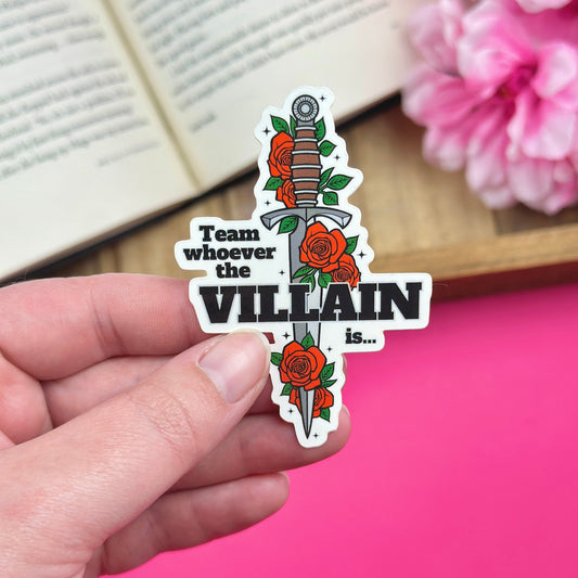 Hand holding a sticker with sword and roses design that says "team whoever the villain is", with an open book and pink flowers in the background.