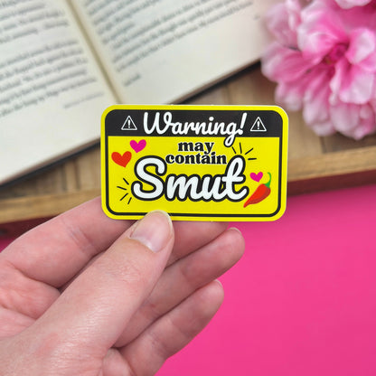 Hand holding a yellow and black vinyl sticker that says "Warning! May contain Smut", with an open book and pink flowers in the background